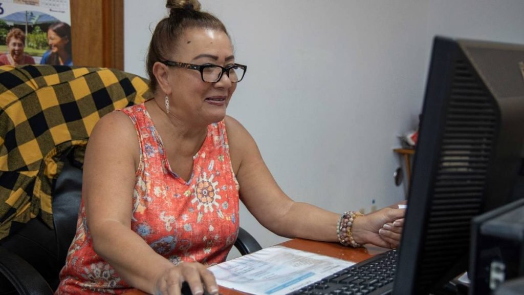Woman with glasses and a bun sits at a desk, typing on a desktop computer.