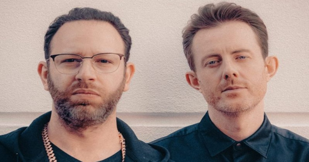 Two men stand side by side against a pale wall, looking at the camera; the man on the left wears glasses and a gold chain, the man on the right has short hair and wears a dark shirt.
