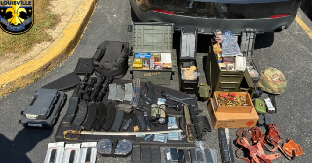Seized weapons and gear laid out on asphalt beside a car, including rifles, magazines, ammo boxes, tactical bags, and police markings in the corner.