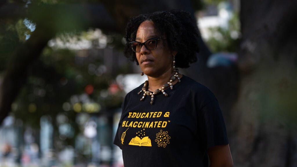 Portrait of a Black woman with short natural hair and glasses, wearing a black T-shirt that reads 'Educated & Blackinated' in yellow with a book icon, outdoors with a blurred background.