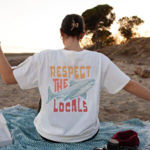 Back view of a person sitting on a patterned blanket at sunset on a beach, wearing a white T-shirt that says 'Respect the Locals' with a blue shark graphic,