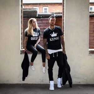 Couple wearing black 'QUEEN' and 'KING' t-shirts, standing with jackets draped over their arms in an urban setting.