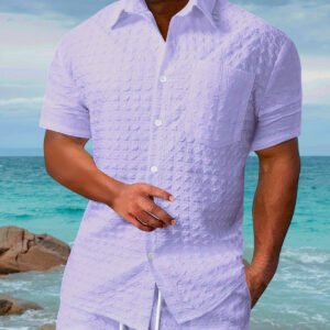 Man wearing a light purple textured short-sleeve shirt standing on a beach with ocean and rocks in the background