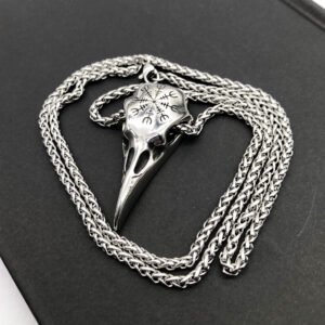 Silver bird-skull pendant with etched runic symbols, resting on a thick silver chain against a dark background.