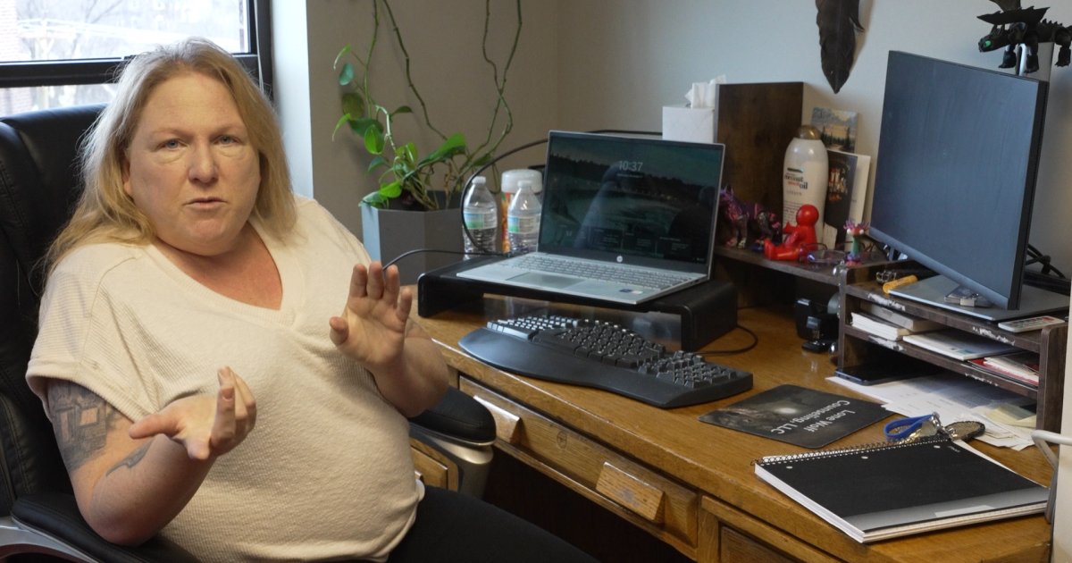 Woman in a white sweater sits at a cluttered desk with a laptop and monitor, gesturing with her hands as she talks in a casual office setting.
