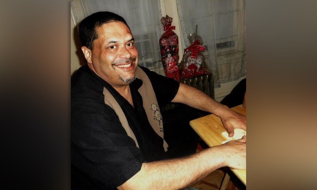 Smiling man with short dark hair and a goatee sits at a table, hands on the wooden surface; Christmas gifts visible in the background behind him.