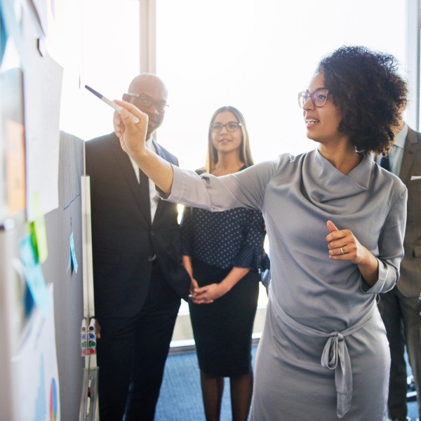 Businesswoman points to a whiteboard with sticky notes while colleagues watch in a modern office.