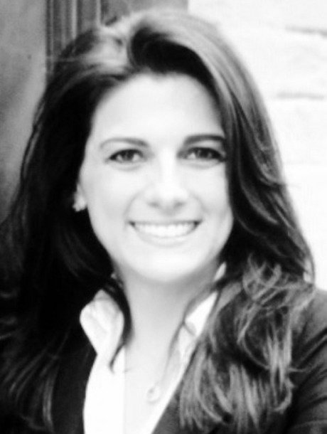 Smiling woman with long dark hair wearing a blazer and white blouse, portrait-style photo in grayscale.