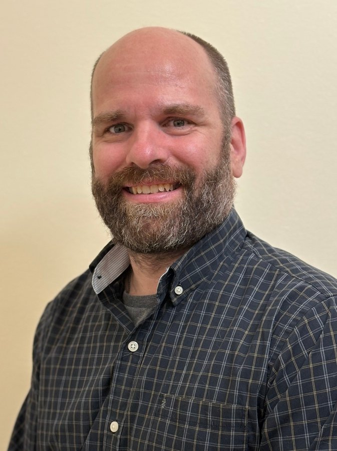 Head-and-shoulders portrait of a smiling, bearded man with a shaved head wearing a dark plaid shirt.