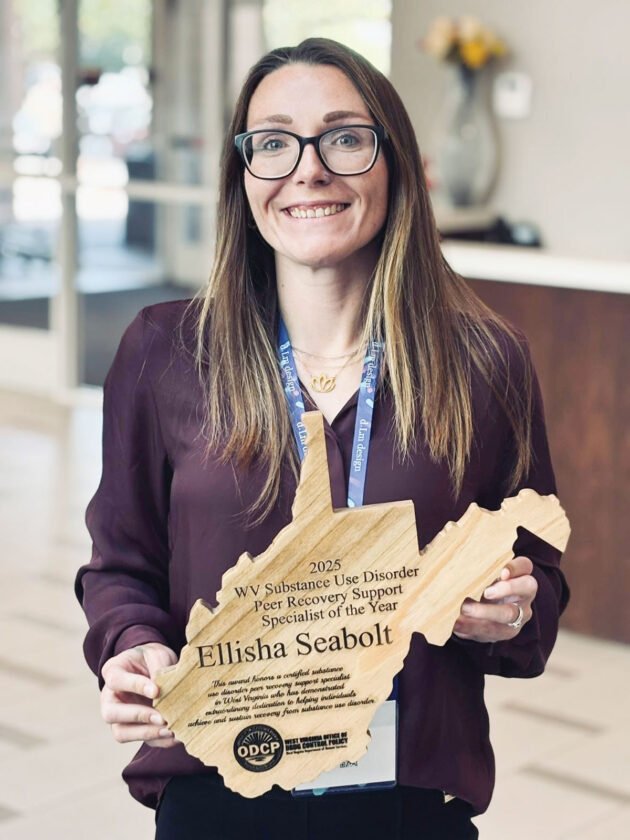 Smiling woman with glasses holding a wooden state-shaped award that reads 'Ellisha Seabolt' for 2025 WV Substance Use Disorder Peer Recovery Support Specialist of the Year
