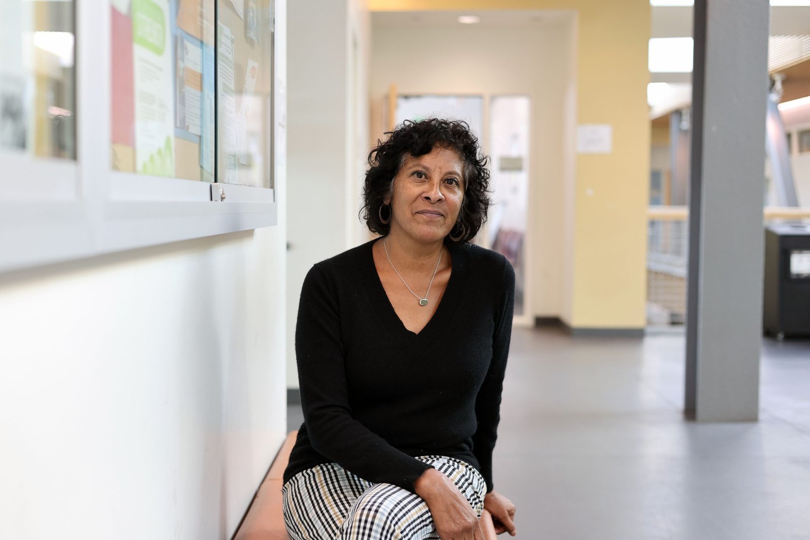 Portrait of a middle-aged woman with short curly hair, wearing a black top and checkered skirt, seated in a bright indoor hallway.