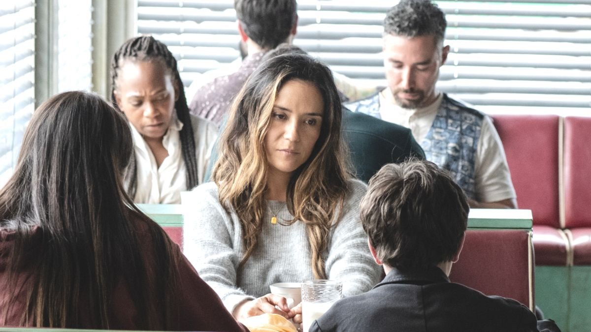 Group of diverse adults seated in a cafe, central woman in a gray sweater looks thoughtful with a cup and glass on the table