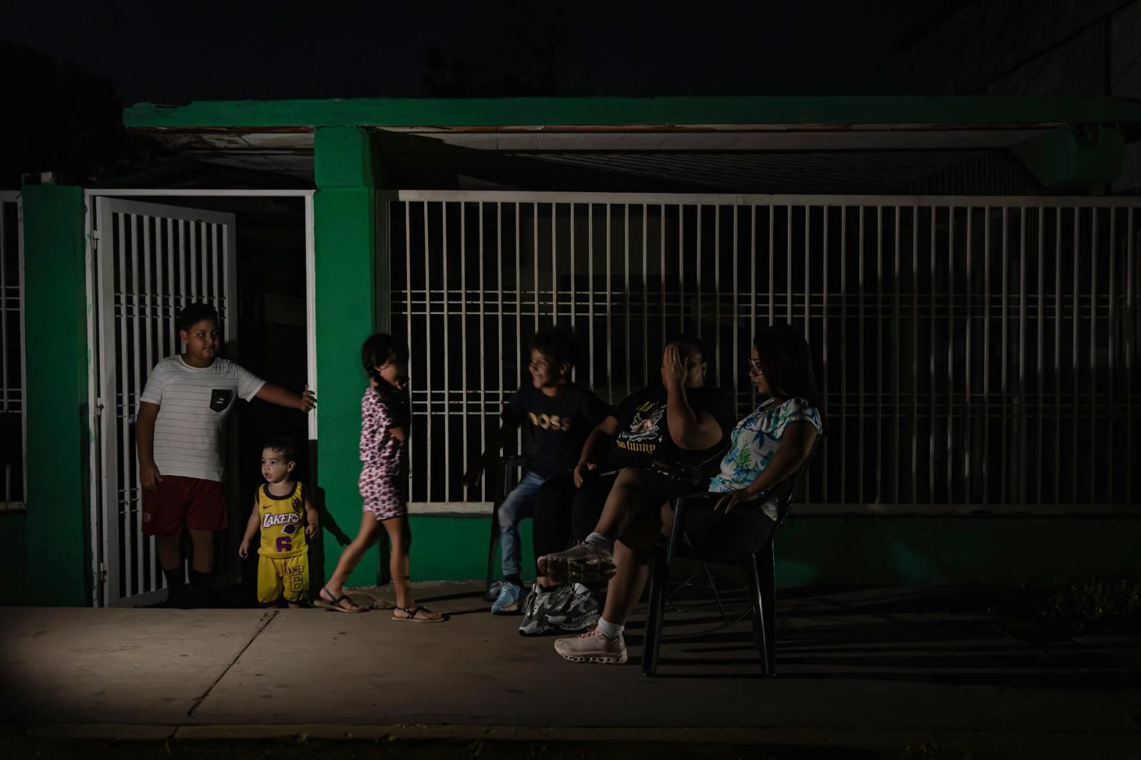 Family standing on a porch at night beside a green building with white metal bars; children and adults sit or stand near the gate.