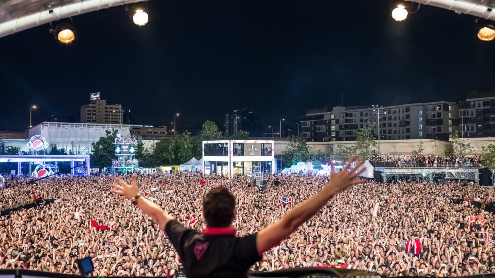 Performer on stage with arms raised facing a packed night-time crowd at a large outdoor concert venue.