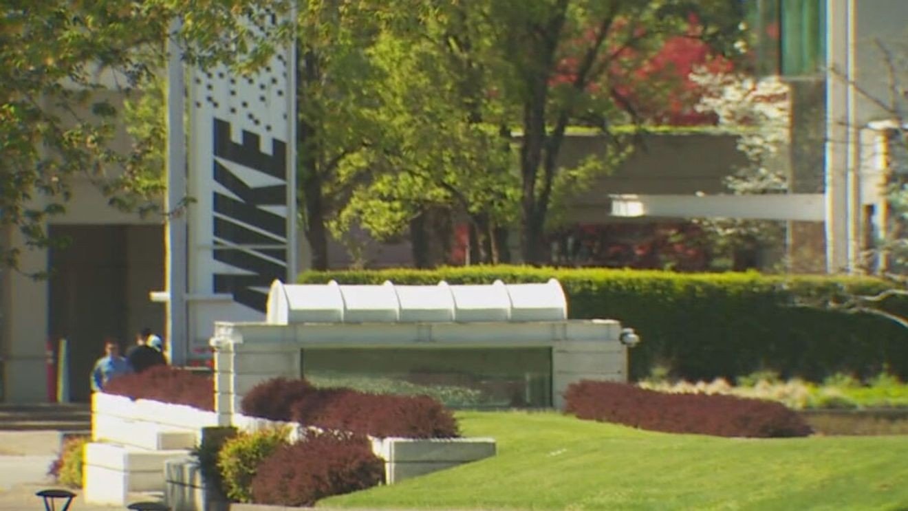 Outdoor campus or office plaza with a white curved canopy over a glass entrance, a trimmed hedge, colorful bushes, and a grassy slope; a tall vertical sign and a couple of people are visible in the background.