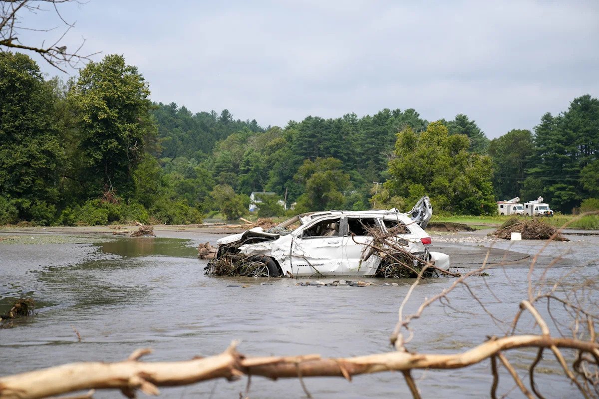 White SUV wrecked and partially submerged in a flooded river, with branches tangled around it and a forested backdrop.