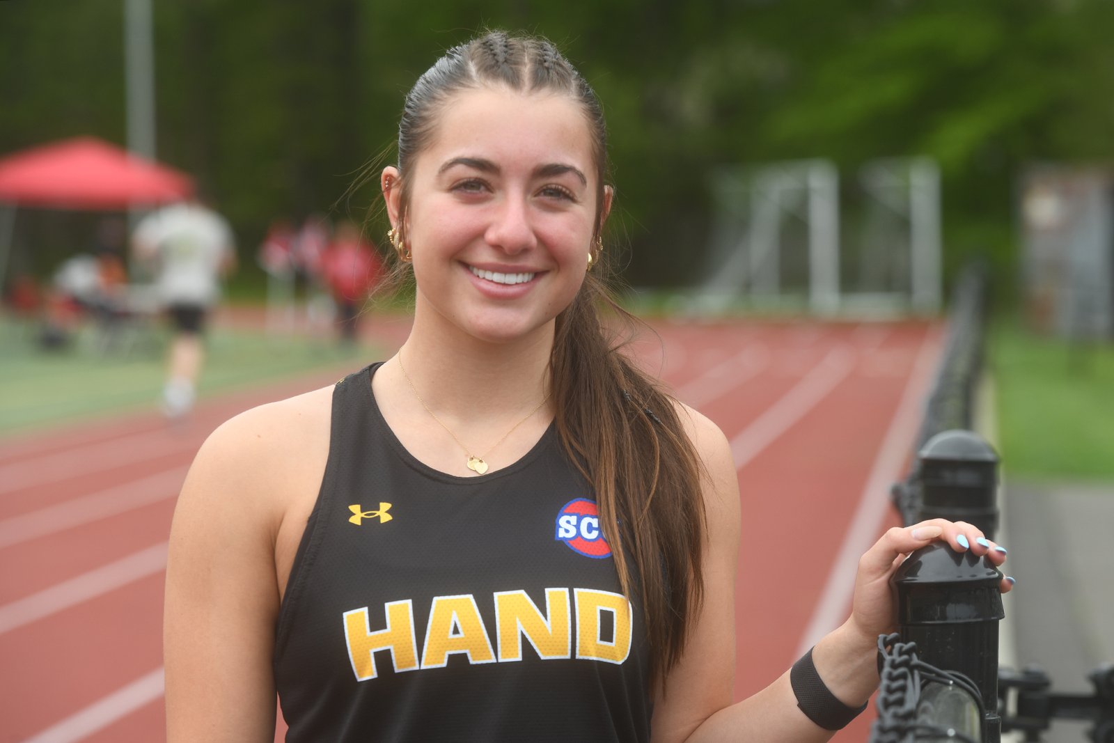 Smiling young female track athlete in a black sleeveless top, standing by a track fence with a water bottle in hand.