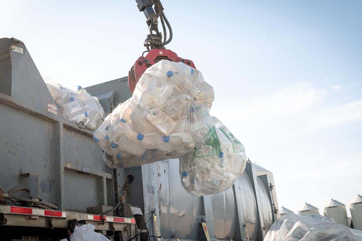 wp-header-logo-656png • Punkin's Patch Large transparent bag filled with plastic bottles being lifted by a red crane attachment into a gray dump truck in an industrial yard.