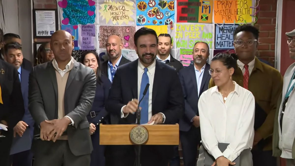 wp-header-logo-692png • Punkin's Patch Man in a dark suit and blue tie speaks at a wooden podium with a microphone, flanked by a diverse group of adults indoors.