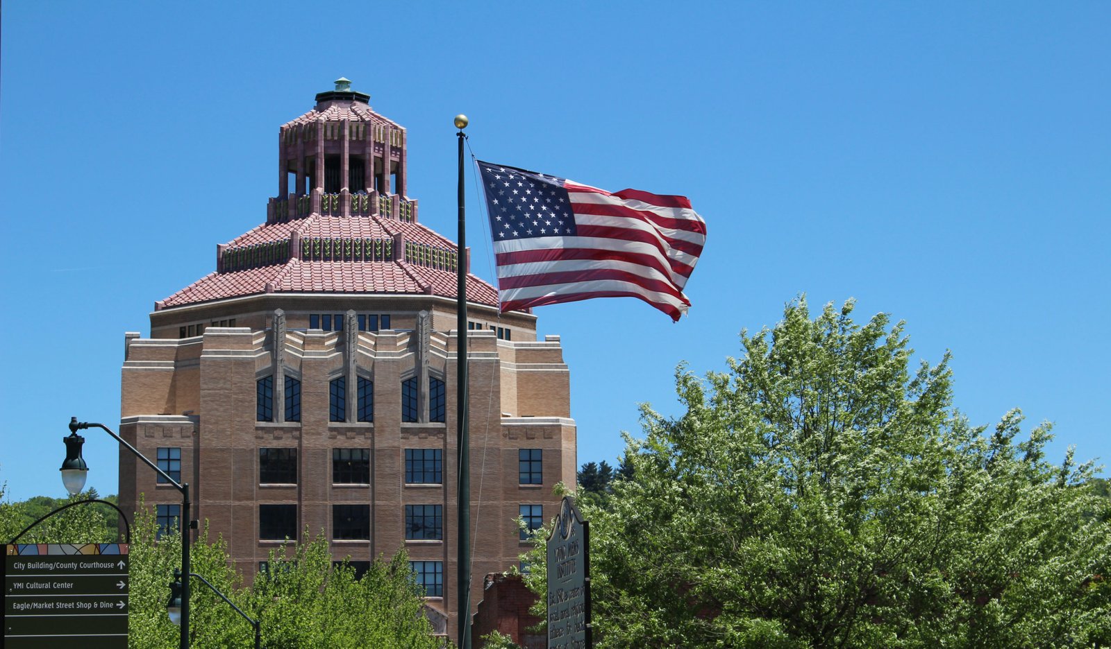 wp-header-logo-693png • Punkin's Patch Brick municipal building with a red-tiled roof and an American flag on a tall pole against a clear blue sky, with green trees to the right.