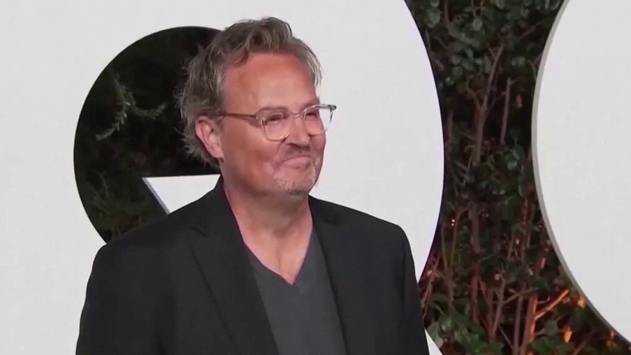 Smiling man with gray hair and glasses wearing a dark blazer and gray shirt at an outdoor event.