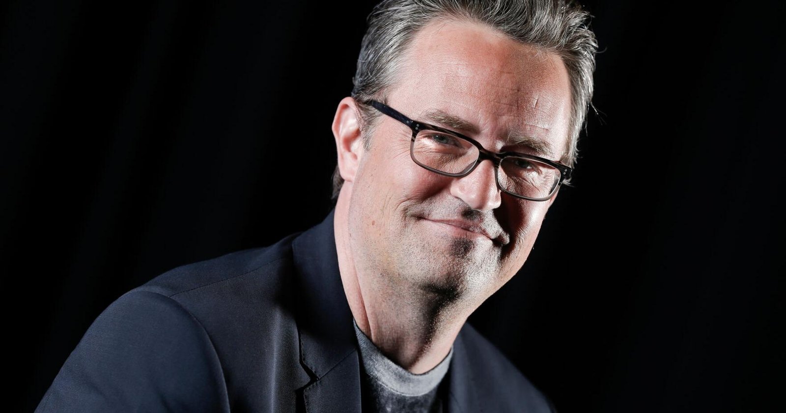 Portrait of a smiling man wearing glasses and a dark blazer, against a black background.