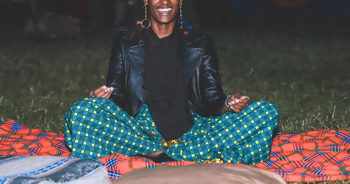Person sitting cross‑legged on a colorful blanket in a grassy outdoor area at night, smiling and meditating with hands resting on knees.",