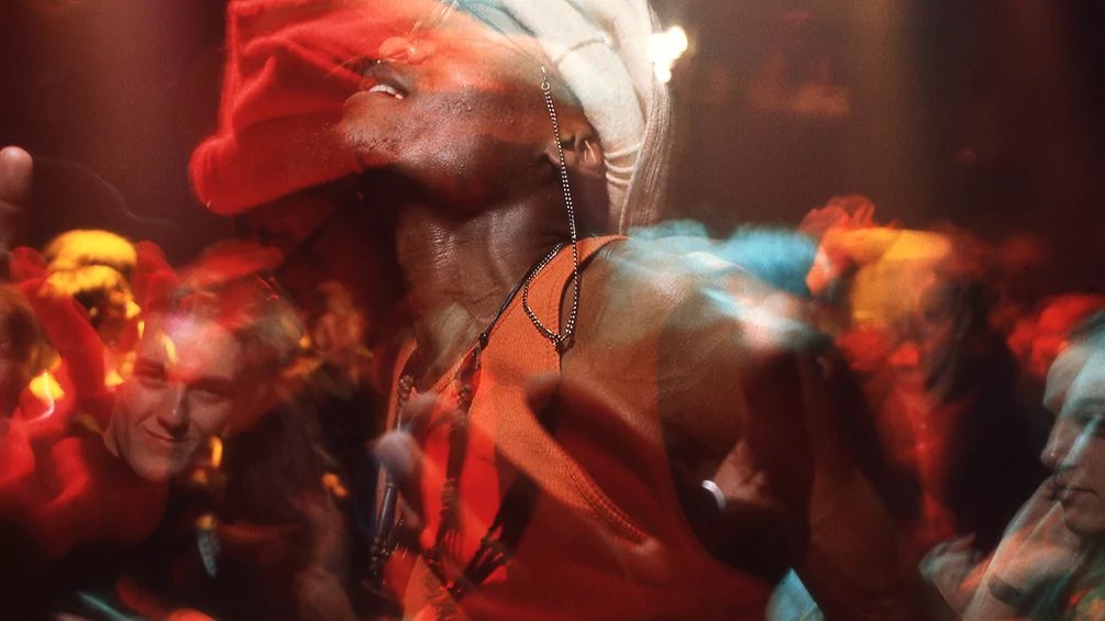 Crowd at a lively concert with a performer in a red head wrap center stage, surrounded by colorful, blurred lights and motion blur in the audience.