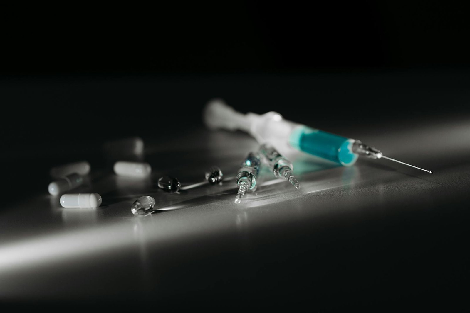 Syringes and assorted pills scattered on a dark surface, suggesting medication or drug use in a dim setting
