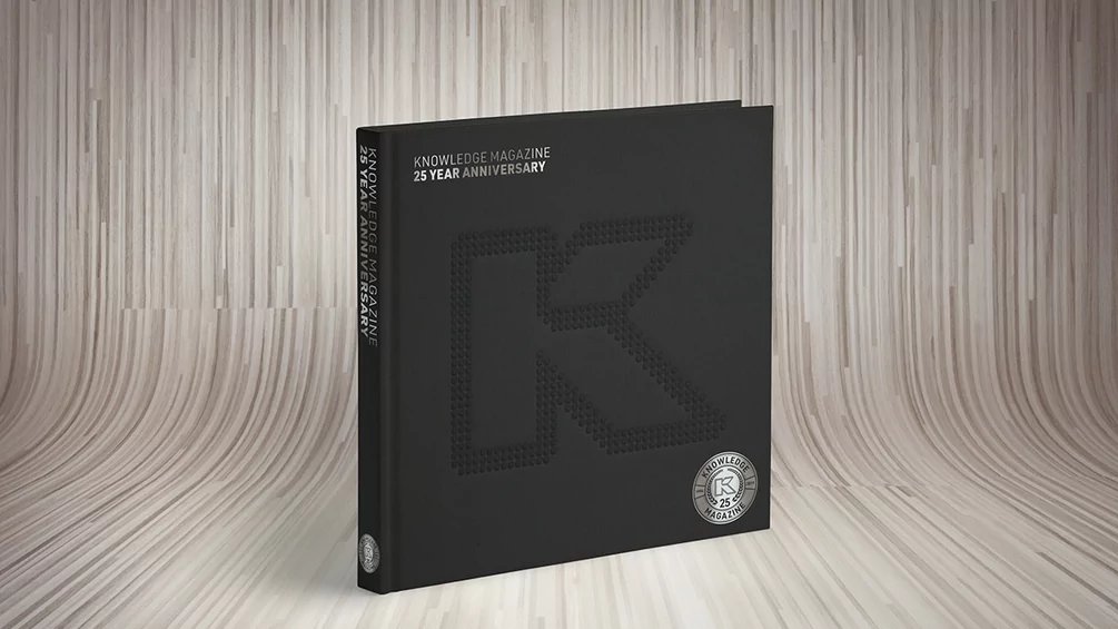 Black Knowledge Magazine 25th Anniversary edition standing upright on a light wood floor, with an embossed K on the cover and a circular seal.