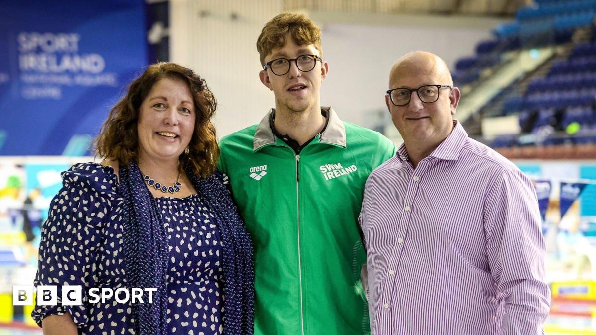 Three people pose together indoors at a swimming venue; the central figure wears a green Swim Ireland jacket, flanked by a woman on the left and a man on the right, BBC Sport logo visible.
