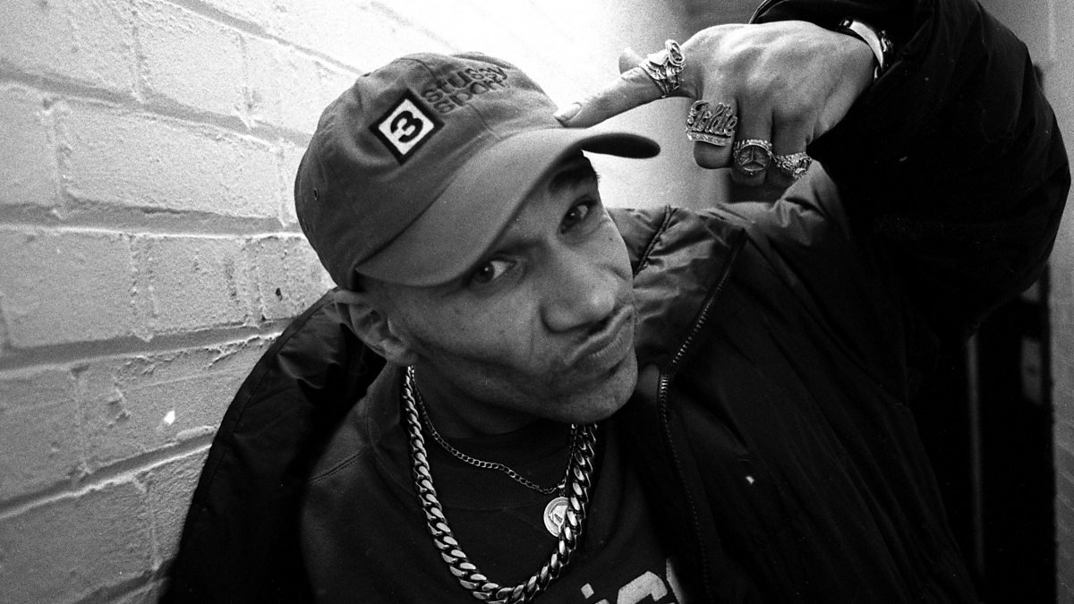 Black-and-white portrait of a man wearing a cap, chunky chains, and multiple rings, leaning against a brick wall and pointing at his cap with a confident expression.