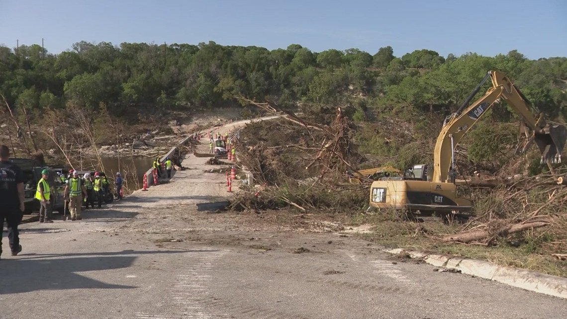 Road blocked by fallen trees and debris; construction equipment and workers repair the area on a damaged rural road in daylight.