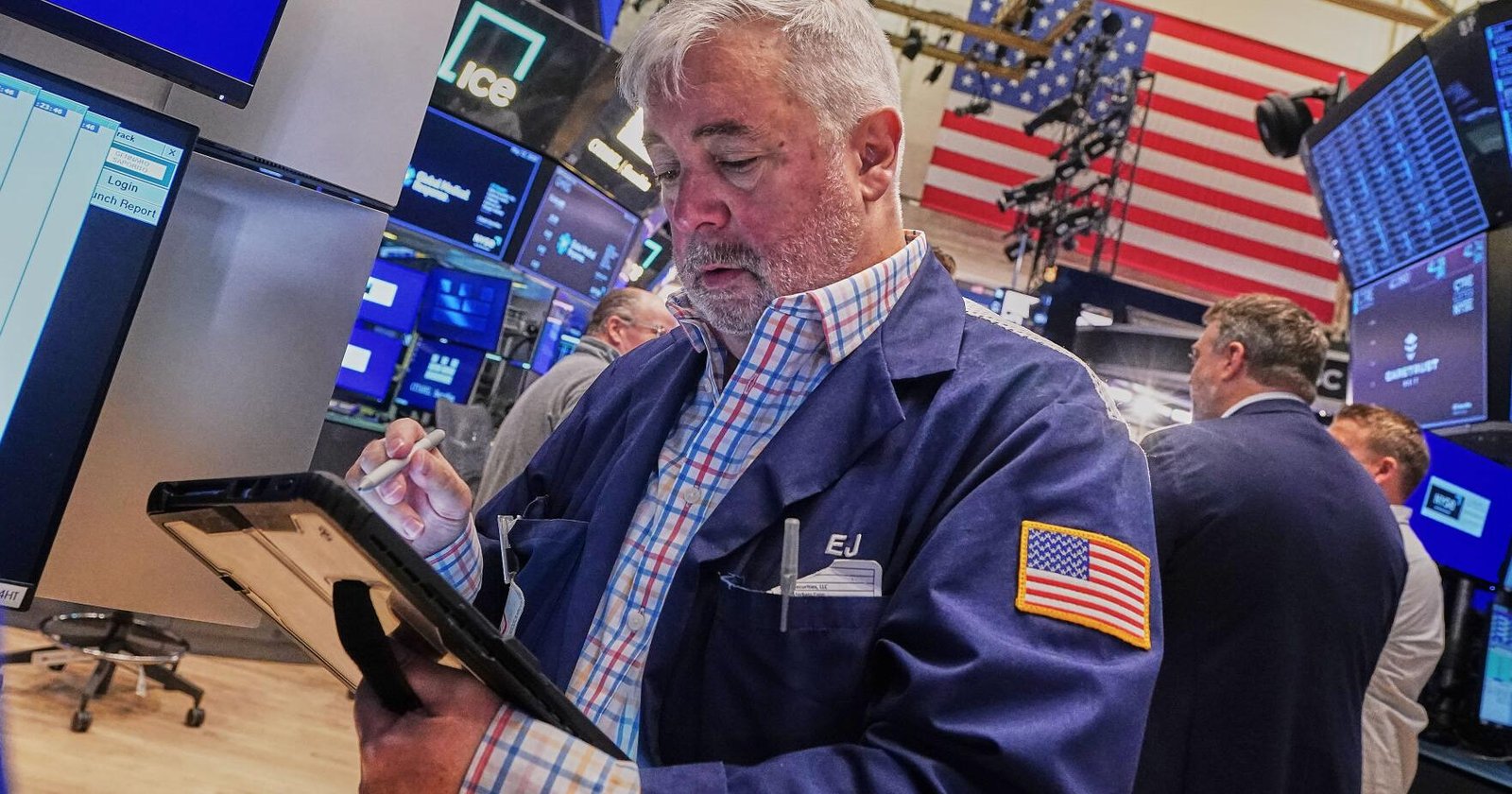 Man in a blue trading jacket with an American flag patch on the sleeve, writing on a tablet with a stylus amid monitors and a busy trading floor