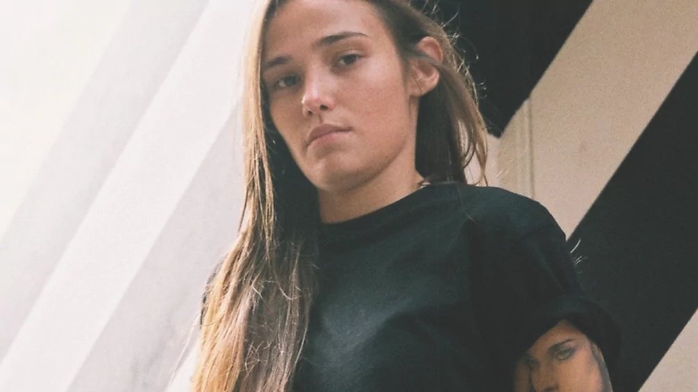 Portrait of a young woman with long hair wearing a black shirt, standing against a striped wall.