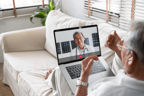 Older man on a video call with a doctor on a laptop screen in a cozy living room setting.