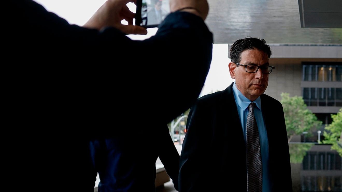 Man in a dark suit and blue shirt walks outside a modern office building while a person in the foreground takes a photo with a smartphone camera.