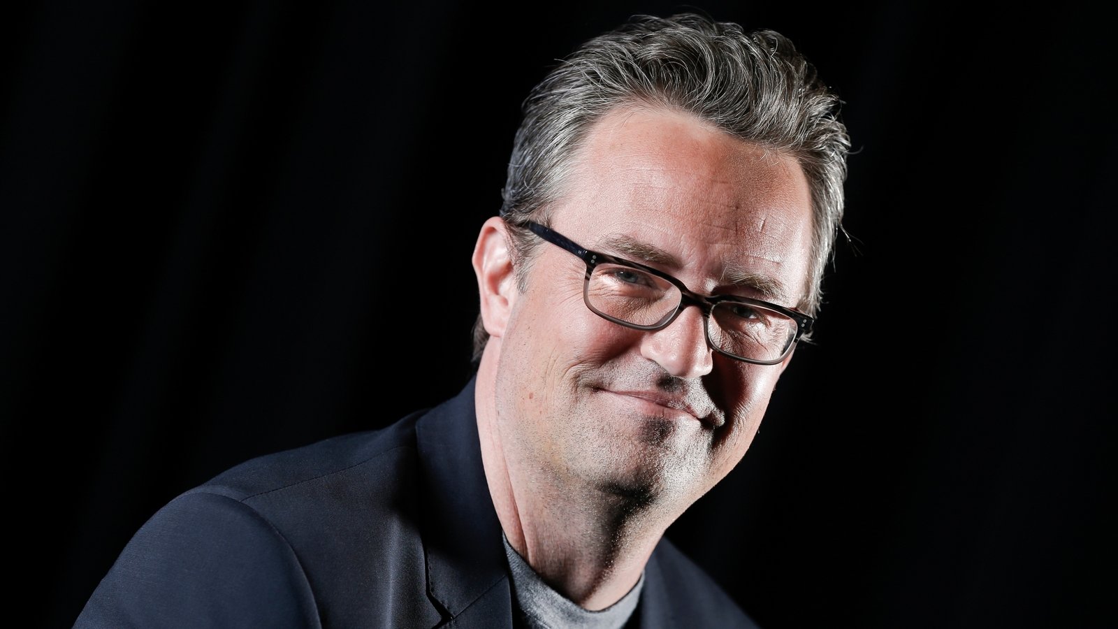 Smiling man with glasses in a dark studio setting, wearing a navy blazer