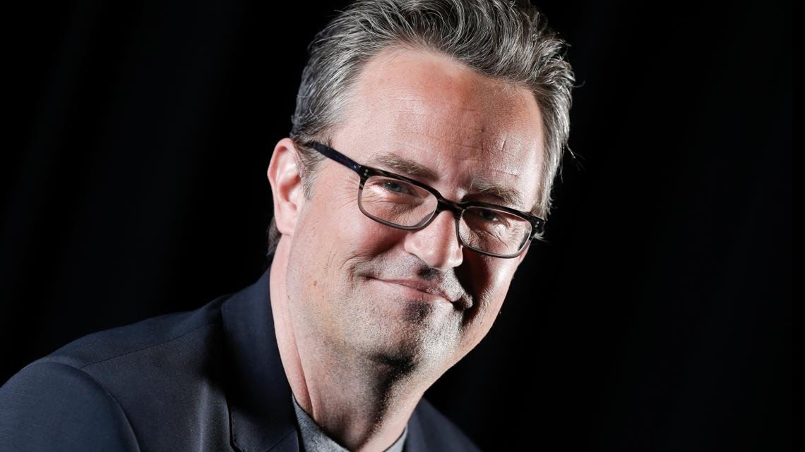 Portrait of a smiling man with gray hair and glasses in a dark blazer against a black background