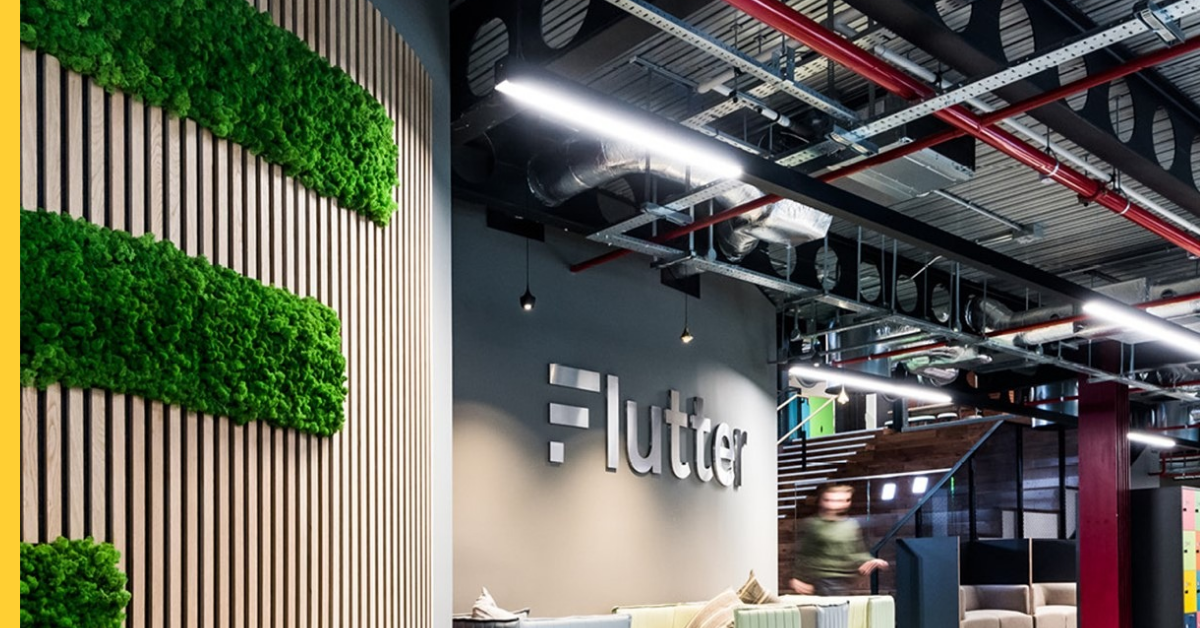 Modern office lobby with a Flutter sign on a gray wall and green moss panels along a vertical wooden slat wall on the left side.