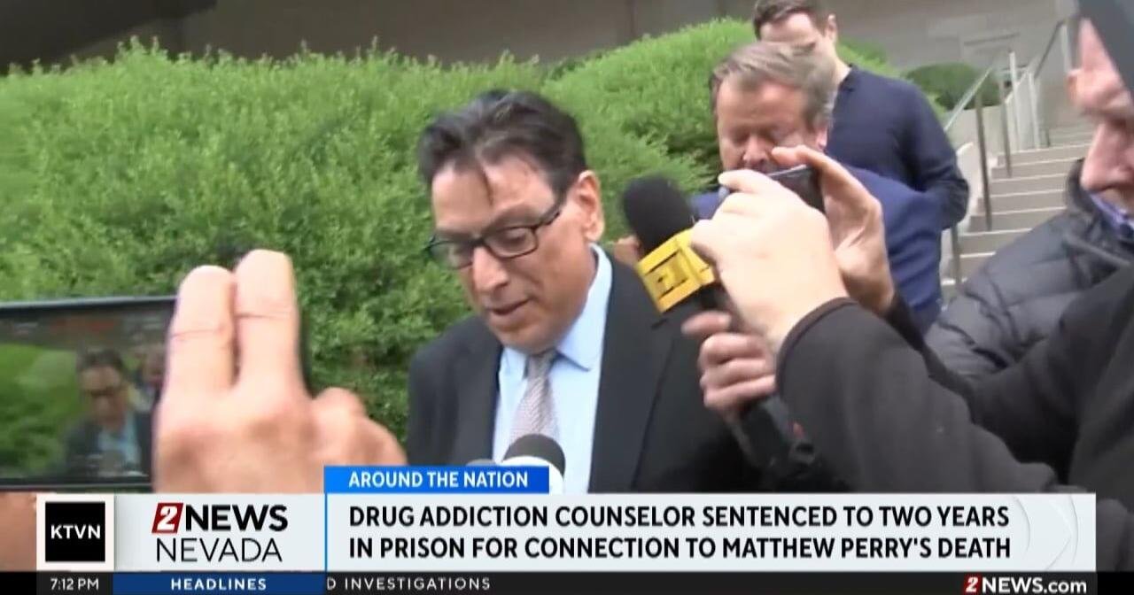 Man in a dark suit and glasses is interviewed by reporters outdoors, with multiple microphones and a TV news banner visible (Nevada / drug counselor sentencing).