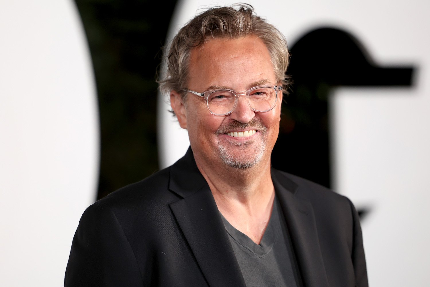 Smiling man with gray hair and clear glasses, wearing a dark blazer over a gray shirt at a formal event.