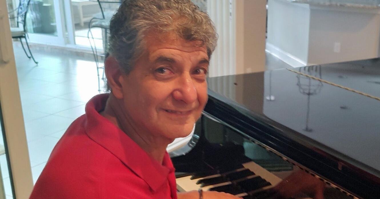 Smiling man with gray hair in a red polo shirt playing a grand piano in a bright room