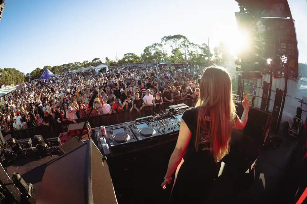 DJ with long hair on stage facing a large outdoor crowd at a festival, mixing on turntables in the foreground.