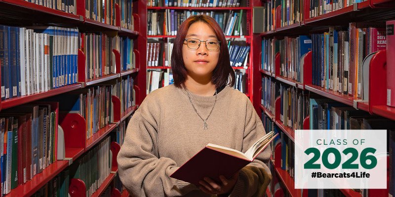 Student with glasses reads a book in a university library, surrounded by shelves; overlay shows Class of 2026 #Bearcats4Life.