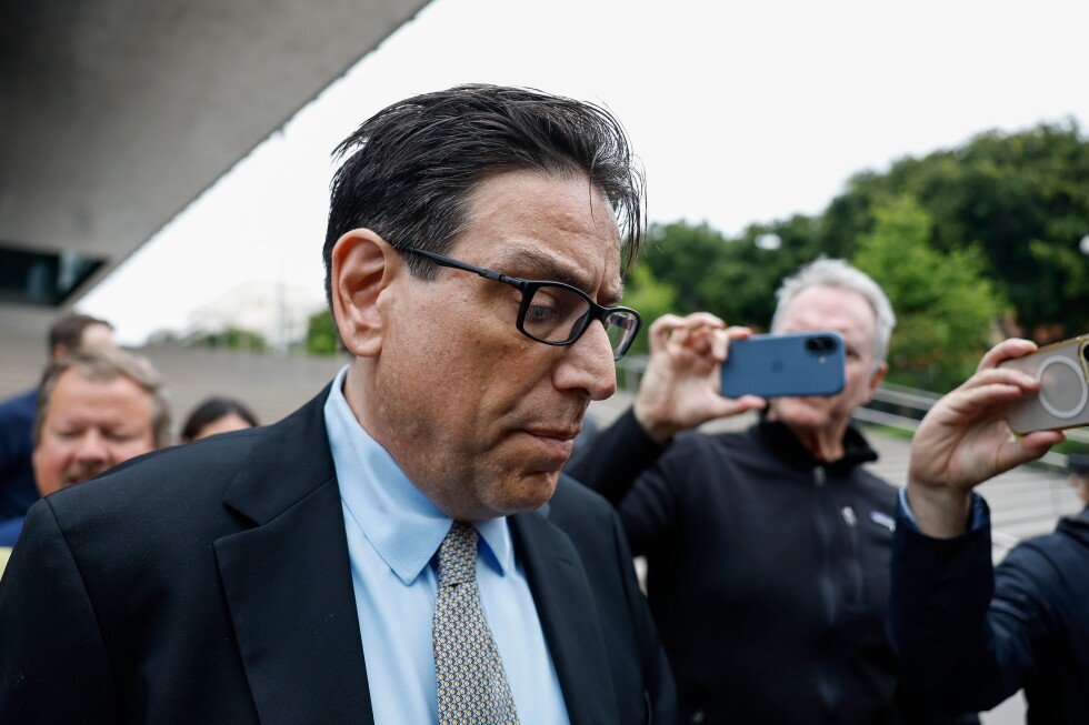 Man in a dark suit and blue shirt looks downward as bystanders film him with smartphones nearby.