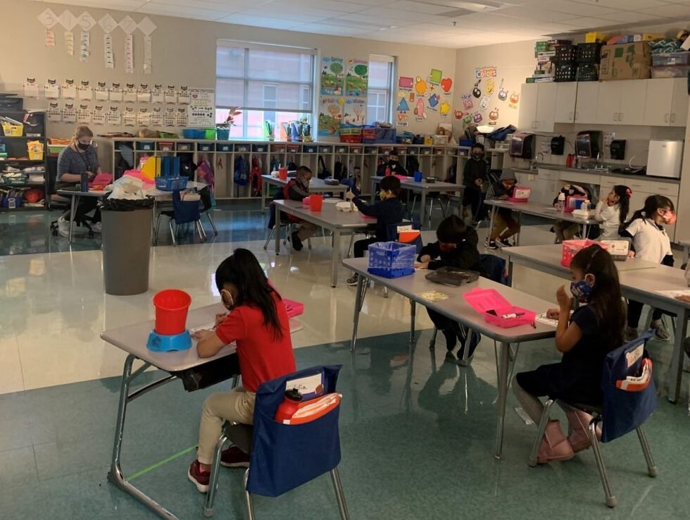 Elementary classroom with students at individual desks wearing masks; teacher sits at left assisting.