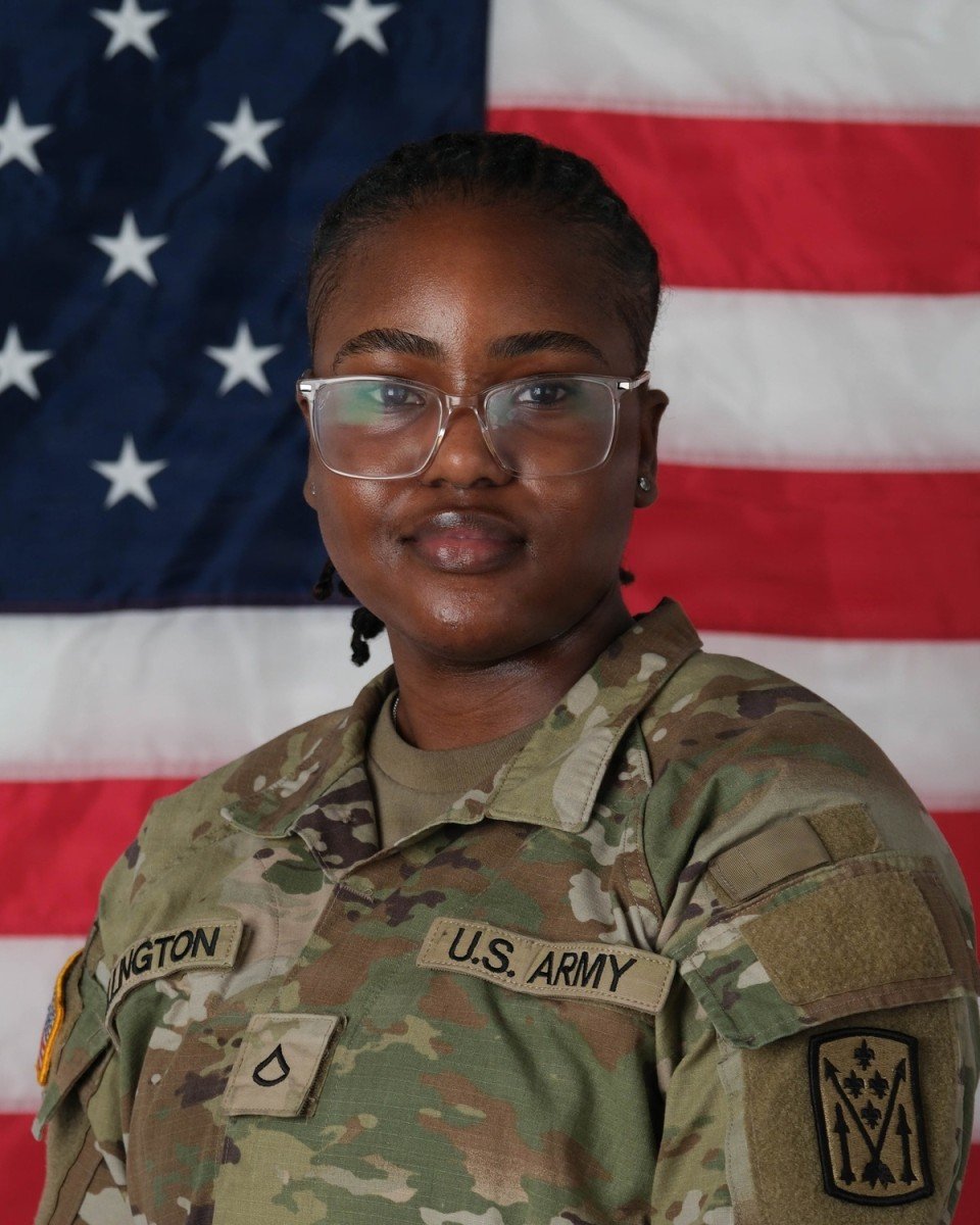 Portrait of a female U.S. Army soldier in camouflage uniform wearing glasses, posed in front of a large American flag.