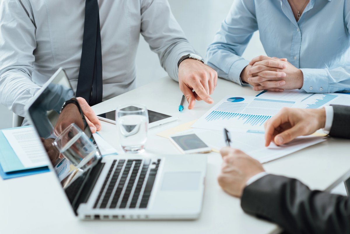 Team of professionals in a meeting reviewing charts and graphs on a white table with laptops and tablets nearby