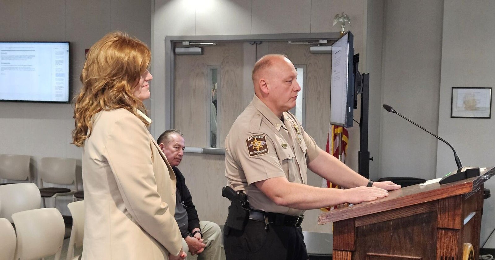 Sheriff in uniform at a wooden podium delivering a speech, with two people seated nearby in beige attire.